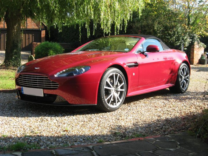 Six Weeks of V8VR S Ownership - Page 1 - Aston Martin - PistonHeads - The image captures a vibrant scene with a sleek red Aston Martin sports car as the centerpiece, parked on a gravel driveway nestled in the sunlight. The car's design is striking, with sharp lines and the luxurious emblem on its grille. The Aston Martin is positioned near a tree and a building with a brick facade, adding an urban touch to the scene. The sunlight casts beautiful shadows and highlights, making the image dynamic and captivating.