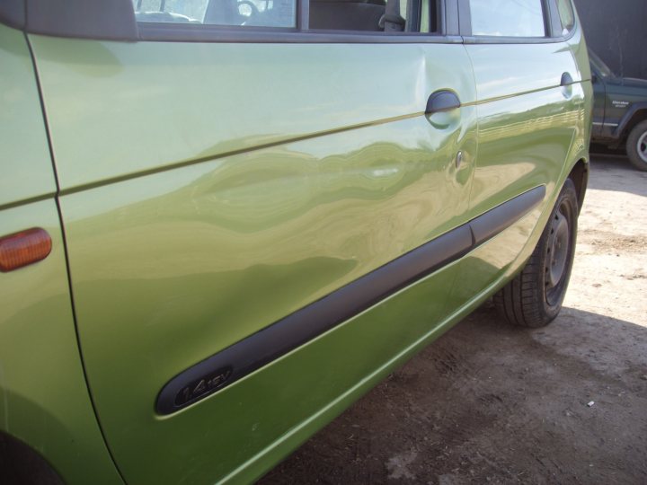 my first car  - Page 1 - Readers' Cars - PistonHeads - The image shows a close-up of a section of a green car bumper. The focus is on the bodywork with a visible slight dent on the left side of the bumper. The car is parked on what appears to be a dirt ground. There is a spare tire visible in the background, indicating a parking area or lot. The overall lighting suggests it could be midday or early afternoon, as the shadows seem soft and natural.