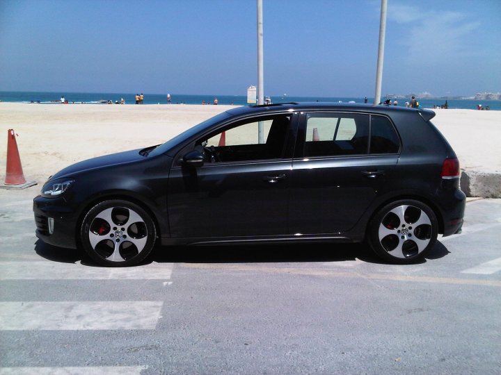 Readers East Pistonheads Drives Middle - The image features a modern black Volkswagen car parked on a street. The car is positioned perpendicular to the curb, with its front facing the camera. The setting appears to be a city street, evident by the road markings visible next to the car. The sky is blue, suggesting a clear day. In the background, there are people and a body of water, possibly an ocean, implying the car might be parked near a beach or coastal area.