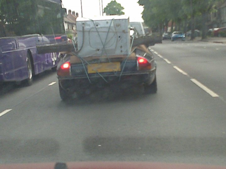 Pistonheads Move Belongings - The image appears to be taken from the passenger side of another vehicle, showing a partial rearview of a car driving on a road. The main focal point is the rear of a light-colored car, which is identified by a yellow license plate. Above the car's rear window, there is a large piece of white machinery or equipment, securely strapped to the vehicle. The headlights of the trailing car are dimly visible under the overcast sky, and there are no visible pedestrians or other types of vehicles in the immediate vicinity. The road itself is two-lane, and the scenery is nondescript, with no distinct landmarks or features visible in the background.