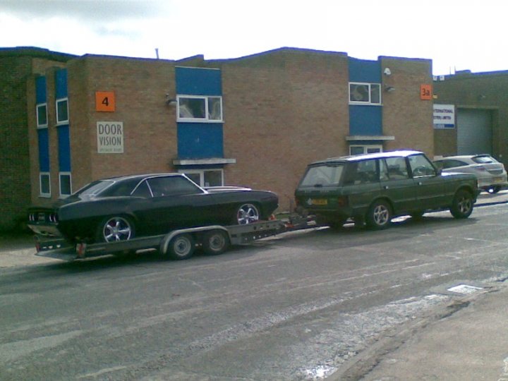 Pistonheads - The image shows a flatbed truck towing a green car on its back. There's a blue and red brick building with the number 4 on the left side of the building. On the right side, there's a white wall and a garage door. The truck is parked on an asphalt road, and the weather appears to be overcast. There doesn't seem to be any activity currently happening in the scene.