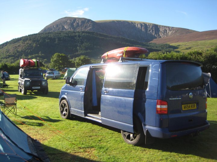 Québec, Canada - Page 1 - USA & Canada - PistonHeads - The image showcases a serene outdoor setting with a blue Volkswagen van parked on a grassy field. The van, a prominent feature in the scene, has its rear door open, suggesting perhaps an ongoing camping activity or preparation for one. Behind the van, a picturesque scene unfolds with a rugged mountain range providing a stunning backdrop. There are other vehicles and tents scattered in the background, indicating that several campers are present in this area. The presence of hikers with backpacks further supports the idea of this being a popular spot for outdoor activities. The overall mood of the image is peaceful and inviting, capturing a moment of tranquility in a world surrounded by nature.