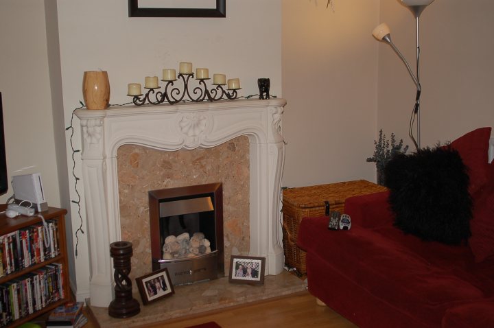 Room Pistonheads Living - The image depicts a cozy living room. A white fireplace, adorned with a row of tiered candles and framed within a silver border, serves as the focal point of the room. To the right, a red sofa is accented with a black throw pillow. Nearby, a wooden entertainment stand holds a Nintendo Wii, and a black and white dog statue adds a touch of whimsy.