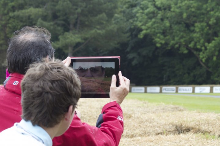 A man and a woman are looking at a cell phone - Pistonheads - The image shows two people in an outdoor setting. One individual is standing, holding a tablet, and appears to be displaying an image. Both people are looking at the tablet screen, which shows a reflection of the person taking the photo. The individuals are wearing casual clothing and the setting suggests it could be a park or a field.