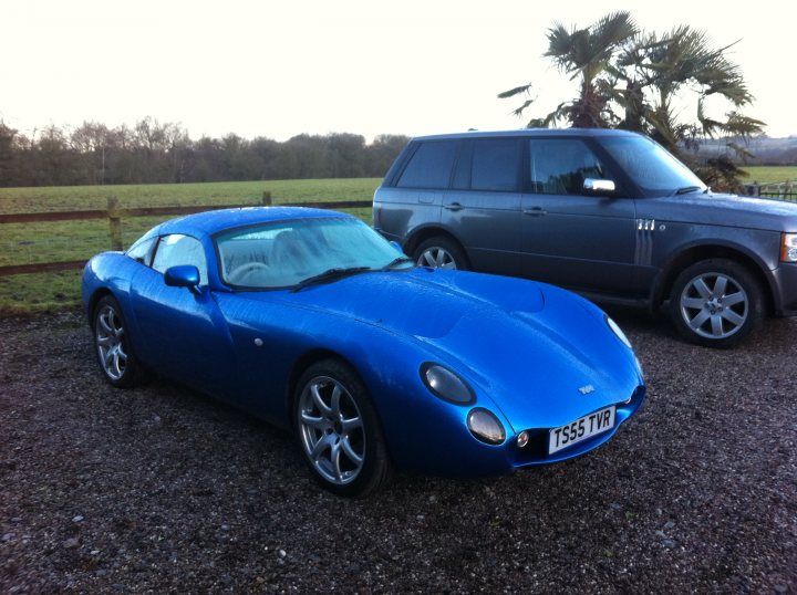 Pistonheads Winter Summer Garage Fantasy - This image shows a blue sports car parked on a gravel surface. The car seems to be a two-door convertible with a hard top. There is a large SUV or crossover vehicle parked next to it, and it appears to be a Range Rover. The surroundings include a fence and some trees, suggesting a somewhat rural setting. The colors and objects suggest that there might be a rainy climate as indicated by the reflection on the sports car's roof. There are no texts or distinguishing marks that are clearly visible in the image.