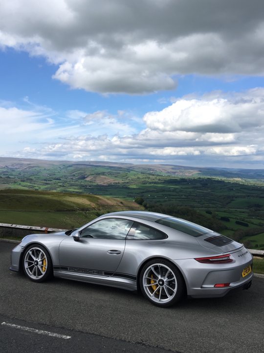 GT3 Touring - Page 37 - 911/Carrera GT - PistonHeads - The image captures a moment on a winding road, where a sleek silver sports car is parked. The car, a convertible, has a striking design and sports an attractive two-tone black and gray roof. The road itself is curved, surrounded by lush greenery, suggesting a rural setting. Above it all, the sky is a vast expanse of blue, dotted with fluffy white clouds. This seemingly quiet moment offers a reflection of the blend of man-made beauty and natural landscape.