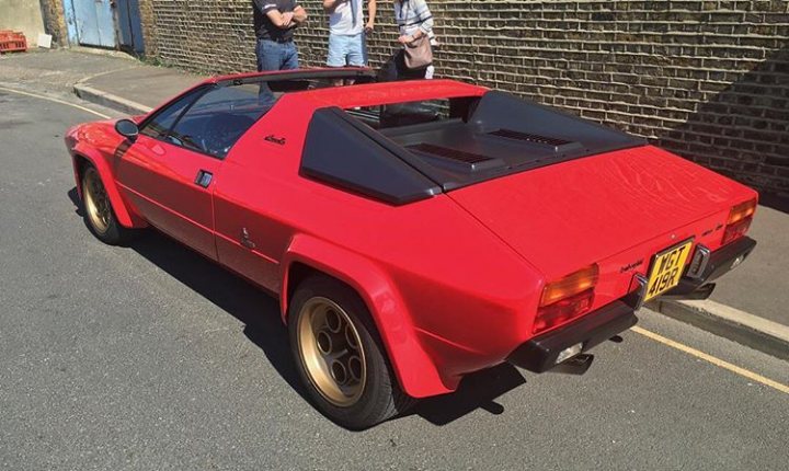 Lamborghini Silhouette  - Page 6 - Lamborghini Classics - PistonHeads - In the image, there's a vibrant red car parked on a narrow street. The car, showing some signs of aging, draws the attention with its dark-tinted rear window and a black roof spoiler. Two people are standing nearby, one of them holding a pink handbag, possibly admiring the brightly-colored vehicle. The car appears to be in a good condition, but the random orange reflector adds a safety element to its design. The street they are on is flanked by a brick wall on one side, suggesting an urban setting, and a sidewalk across the way, where one might see more pedestrians.