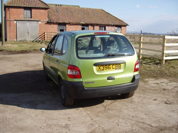 my first car  - Page 1 - Readers' Cars - PistonHeads - The image presents a green hatchback car parked in a dirt area. The car has a contrasting black rear bumper. It is facing towards the right side of the image. There is a brick and tile building in the background, suggesting a rural or semi-rural setting. A wooden fence can be seen to the right of the car. The sky appears to be clear, and there are clouds visible above the building.