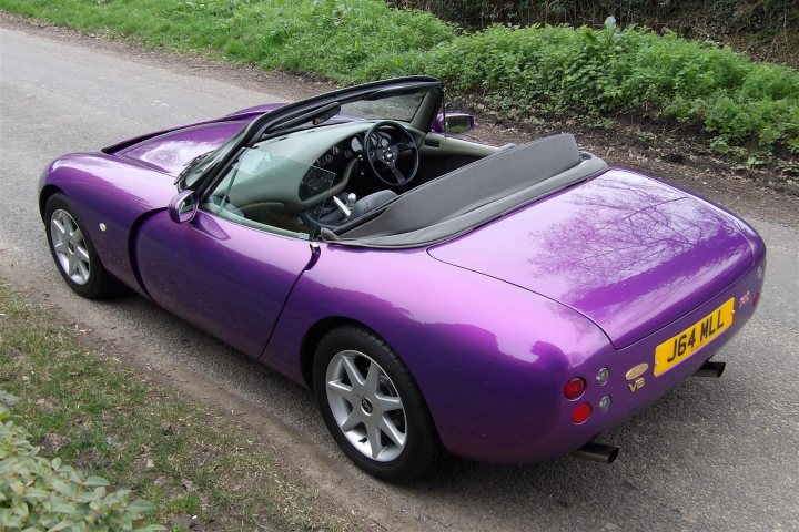 One of each - Page 1 - Griffith - PistonHeads - The image showcases a vibrant purple sports car parked on the side of a road. The car's top is down, revealing its sleek interior and black leather seats. It's positioned on a gravel shoulder, adjacent to grass. The surroundings appear to be in the outskirts of a town or village, indicated by the presence of a wooden fence visible in the background.