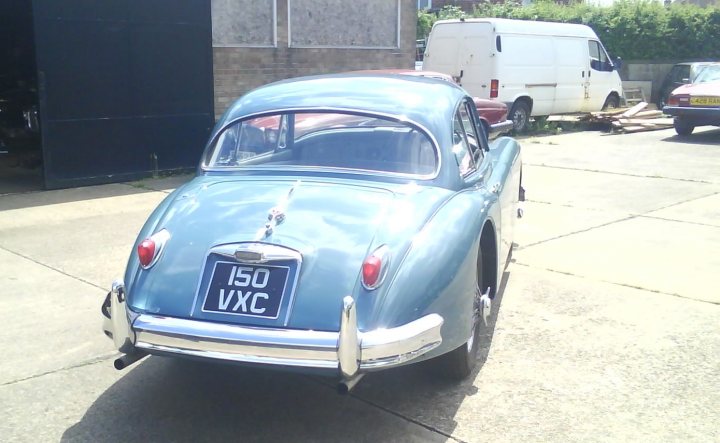 Pistonheads Campbells Donald Etype - This image shows a vintage car with a unique blue and silver color scheme, identified as a Vauxhall model, parked in a driveway. In the background, there are other vehicles, including a red car and a white van, as well as a darker colored vehicle whose make is not clearly visible. The setting appears to be an urban or semi-urban outdoor area, with paved ground and buildings, suggesting a residential or commercial area. The lighting in the photo is bright, possibly indicating it may be a sunny day.