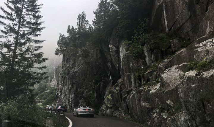 Road trip of a lifetime - Page 1 - 911/Carrera GT - PistonHeads - The image captures a winding road carving its way through a lush, mountainous landscape. The road is surrounded by tall, evergreen trees that add a sense of depth and isolation to the scene. The weather appears foggy, giving the landscape an ethereal, misty quality. Multiple vehicles, perhaps including a tour bus, are traveling along the same road, hinting at some level of human activity in this natural setting. The overall scene conveys a sense of adventure and tranquility in the midst of the natural world.