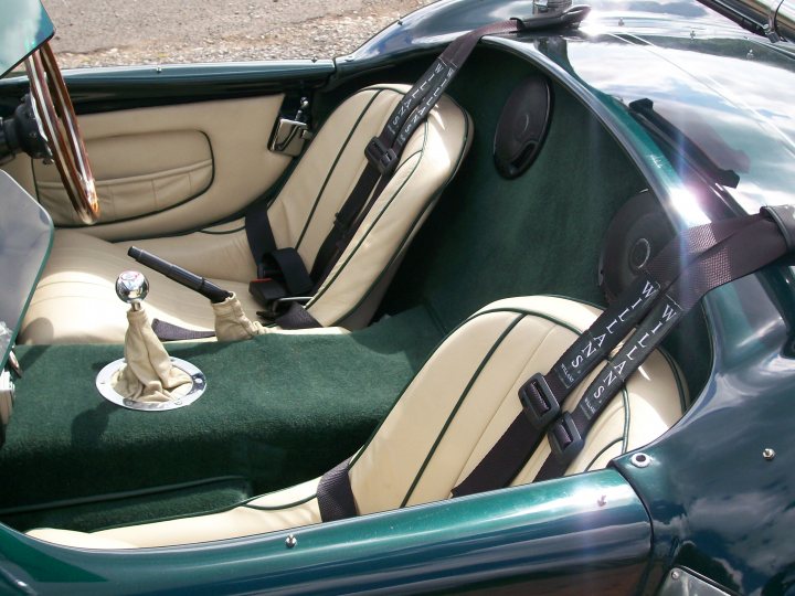 Interior Pistonheads - This image captures the interior of a convertible sports car. The car features a two-seat configuration with a black leather interior and green accents. A silver steering wheel, accompanied by a matching hood ornament, is visible in the foreground. The car is parked on asphalt, and the sunlight streaming in suggests an early morning or late afternoon setting. The overall atmosphere of the image is sleek and luxurious.