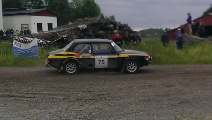 SAAB 99T rallycar pics - Page 1 - Saab - PistonHeads - The image captures an exciting scene set on a dirt track. At the center of the action, a small black and yellow car, emblazoned with the number 75, is tearing through the track, its ability evident from the dust it's kicking up. A group of spectators are visible in the background, their attention focused intently on the car, some even using their cell phones to capture the thrilling moment. On the sidelines, damaged trees can be seen, possibly caused by a fallen power line, adding an element of danger to the scene.