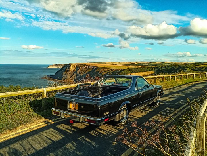 Lets see your Yank tanks! - Page 53 - Yank Motors - PistonHeads UK - The image shows a classic muscle car, specifically a dark-colored Chrysler with a long hood parked on the side of a road. The car has an open trunk and is situated in what appears to be a coastal or rural area with a cliffside or hillside location, as evidenced by the ocean view in the background. The sky is partly cloudy, suggesting fair weather conditions. In the foreground, there's a fence and a tree on the left side of the frame. The perspective of the photo suggests it might have been taken with a wide-angle lens or from a slightly elevated angle.