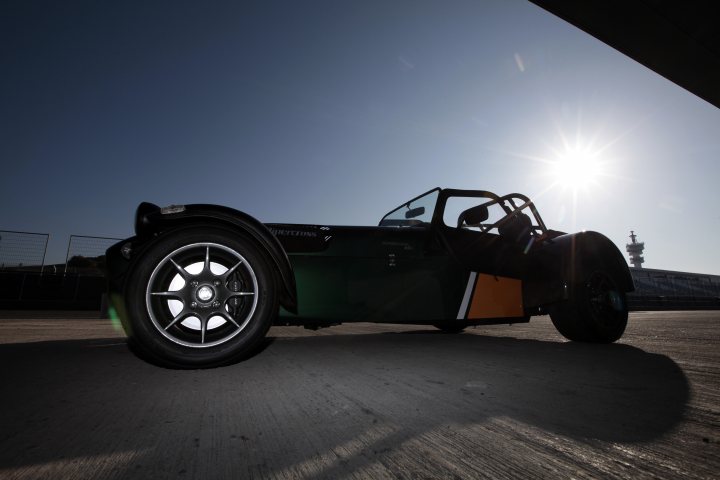 Some Pictures of my R500 Signed - Page 1 - Caterham - PistonHeads - The image captures a vibrant scene of a dark green sports car parked on an airfield. The sun, although low in the sky, is casting a bright light, creating a play of shadow and light on the car. There's a sense of solitude and speed suggested by the empty runway and the powerful stance of the vehicle. On the left side of the runway, there's a single fence, adding to the aviation-themed setting. The overall mood of the image is dynamic and energetic, with the sunset adding a touch of warmth to the otherwise cool colors of the car and the runway.