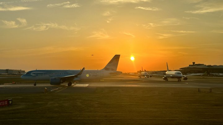 The image presents a tranquil scene at an airport during sunset. The sky is ablaze with hues of orange, casting a warm glow on the airplanes parked on the runway. Two large airplanes are visible in the background; one has a blue tail while the other features a white tail. A few smaller planes can be seen near them. The sun's rays illuminate the scene, creating a serene atmosphere. There is no text or distinctive objects that stand out. This is a commonplace image depicting a typical end of day at an airport.
