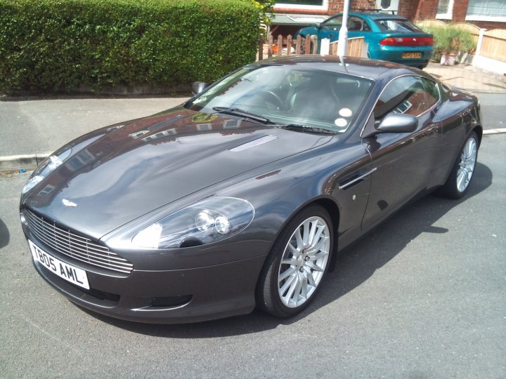 Pistonheads - The image features a silver Aston Martin sports car parked on the side of a street. The car is sleek and shiny, suggesting it is well-maintained. The style of the car is sporty and suggests speed and power. The car is parked in front of a house. There are other cars visible on the street, but they are not as prominent in the image. The focus is clearly on the Aston Martin.