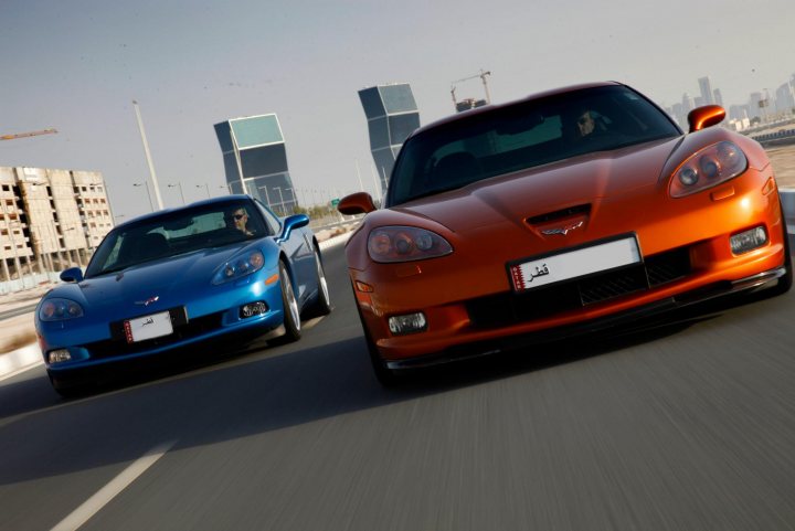 The first Qatar Pistonheads gathering (pics) - Page 1 - Middle East - PistonHeads - This image depicts two luxury sports cars on a highway, captured in motion. The vehicles appear to be blue and orange Corvettes, setting against a backdrop of city buildings. The photographer has captured the high-speed action, as indicated by the vehicle's motion blur. The perspective places the viewer as a passenger in the driver's seat of one of the vehicles. There are no visible texts or notable landmarks. The image evokes a sense of speed and competition.