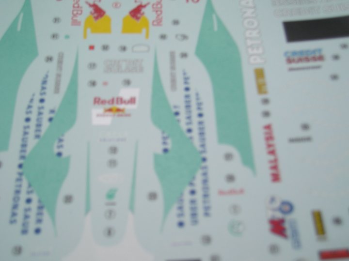 Sauber Petronas C22: 1/24 Revell G.B - Page 1 - Scale Models - PistonHeads - The image shows a close-up of a sticker, which is partially peeled off. The sticker features a colorful design with blue and black elements, including what appears to be a human figure, animal figures, and text written in a mix of uppercase and cursive fonts. The sticker includes the Red Bull logo, indicating a corporate brand identity. Some of the words are obscured, but the sticker also contains elements related to math and sports. The style of the image is clear and the focus is sharp, allowing the viewer to clearly see the details of the sticker.