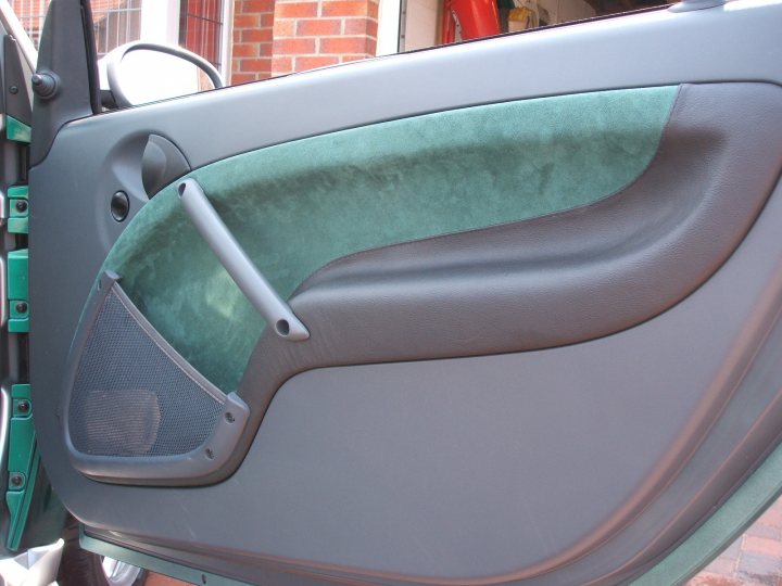 Pistonheads Upholterers - The image features the design of the door and window of an automobile, likely the back of a seat or cargo area. A black and green armrest with a contoured shape is visible, highlighting the considered ergonomics of car interior design. The seat is equipped with a cup holder on the side, under the armrest, indicating the utility of the space for storage. The focus is on practicality and comfort, with attention to detail in both aesthetics and function. In the background, a hint of a red wall provides a color contrast, emphasizing the form and function of the car seat's design.