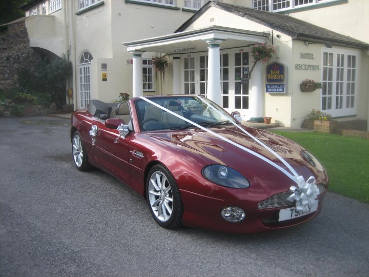 Pistonheads - The image shows a modern red sports car parked in front of a traditional-style building with bay windows. The car features a shiny finish and a convertible top that is folded down, revealing a white interior. The building has a welcoming atmosphere, with a sign indicating it's a reception area, possibly for an establishment. The car is adorned with a ribbon and a decorative lapel pin, suggesting that the vehicle is part of a special event or ceremony. The overall setting is bright and clear, with ample light reflecting off the vehicle's surface.