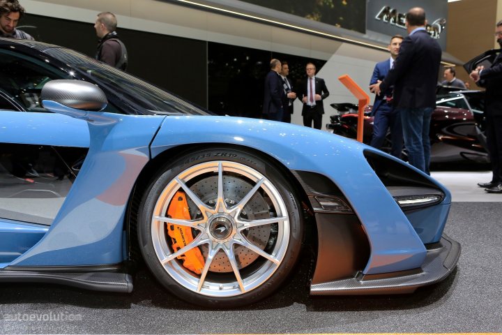 Senna at Geneva - Page 1 - McLaren - PistonHeads - The image depicts a public exhibition showcasing automobiles. The central focus is a blue race car with its hood open, allowing a clear view of the orange wheels and internal mechanisms. Several people are present in the scene, standing around the car, some of whom appear to be inspecting the vehicles. In the background, there's another red car partially visible and another person in the foreground. The setting suggests an auto show, with an array of cars on display for visitors to appreciate and learn more about.