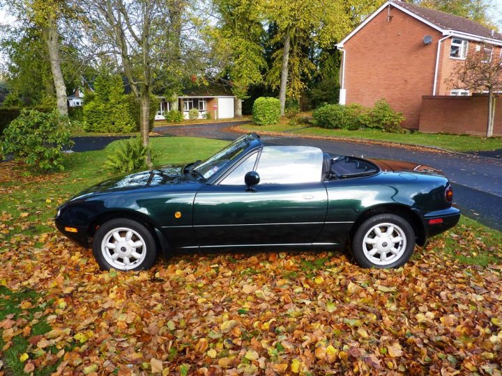 Does a rust free mk1 mx5 exist for £1500 ? - Page 1 - Mazda MX5/Eunos/Miata - PistonHeads - The image displays a dark green convertible sports car parked on a lawn covered with fallen leaves. The car is in a residential area, and behind it, there's a driveway leading to a house. The scene is bathed in natural light, hinting at either dawn or dusk. The car features a convertible top that is up, and there is no visible damage or noticeable branding in the image.