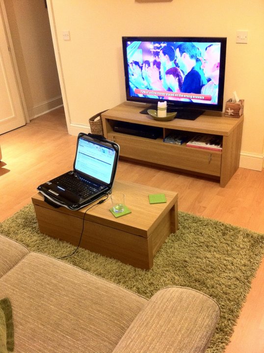 Pistonheads - This image presents a cozy living room setting. A flat screen TV is prominently displayed against one wall, its screen filled with a vibrant array of colors indicating active viewing. Across the room, a dining table occupies the space, on which rests a laptop, a testament to a blend of modern technology and home comfort. Below this table, a rug adds warmth to the rustic ambiance, with its fringed edges adding a touch of homeliness. The room, adorned with wooden furniture, is brightly lit, enhancing the contrast between the cool tones of the wall paint and the warm tones of the wooden surfaces.