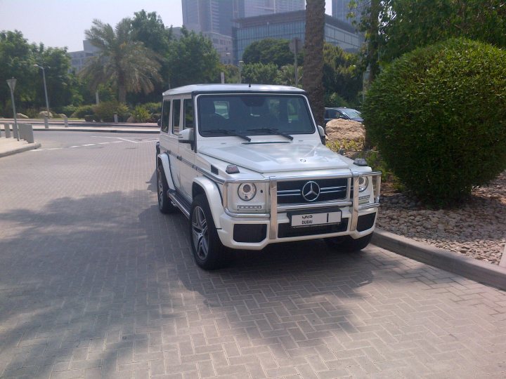 Middle East spotted thread - Page 57 - Middle East - PistonHeads - The image depicts a white Mercedes-Benz SUV parked on a brick street next to a curb. The car has a sleek design with off-road tires and is set against a backdrop of a cityscape including trees and a large building in the distance. In the foreground, there's a partially visible white pedestrian sign that is angled towards the camera. There's no text or distinctive branding visible in the image. The vehicle is situated in a neighborhood with paved roads, and the overall lighting suggests it could be either morning or late afternoon.