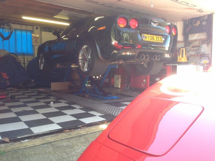 Z06 day today - Page 1 - Corvettes - PistonHeads - The image showcases a garage with a car hoisted on a hydraulic lift. The car appears to be a sports or luxury model, with a distinctive appearance, possibly hinting at a convertible body type. The garage floor is painted with black and white checkered squares, indicating a sturdy and durable surface suitable for vehicle maintenance. Various tools and equipment are scattered around the garage, suggesting that this is a well-used and active workspace. Lastly, the garage's walls and ceiling are filled with various items, including shelves and racks, indicating a space designed for both work and storage.