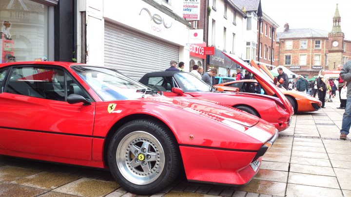 Ormskirk MotorFest - Page 1 - North West - PistonHeads - The image captures a vibrant street scene dominated by a row of red Ferrari sports cars parked on the sidewalk. The Fiennes Art dealership is situated on the corner of the street, adding a touch of elegance to the urban setting. The presence of passersby and beauty shops suggests a busy, lively atmosphere. The brick road beneath the cars and the surrounding buildings further emphasize the urban environment.