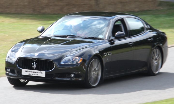 What's the ultimate "family" car? - Page 1 - General Gassing - PistonHeads - The image showcases a black Maserati car driving down a road. The car is in motion, captured in the lower half of the image. It has four doors and a distinctively grilled front. The rear of the car is visible, and the Maserati logo is prominent, confirming the brand. The road appears to be a busy highway, and the vehicle's license plate reads "QUALITY." There is a single individual in the background, partially visible, adding context to the image.