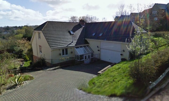 Pistonheads - This image shows a flat sides two-story house under sunlight, with a white garage attached. The house has a distinctive rounded roof and grey paneling. A large bright window allows for ample natural light inside. The driveway is adjacent to the building and leads up to a double garage door. Grass is visible towards the front and sides of the property, suggesting a well-maintained garden area. The sky is partly cloudy, which adds a dynamic element to the scene.