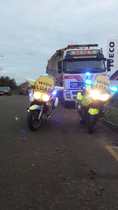 Abnormal load affecting roads this Saturday - Page 1 - Thames Valley & Surrey - PistonHeads - In the image, a wide load is being transported down a street on a cloudy day. Leading the convoy is a white truck with a yellow caution sign, and two police motorcycles are ridden ahead of the truck. The motorcycles have a siren light flashing, warning other vehicles to watch out for the oversized load. The caution sign on the truck also reads "WIDE LOAD," further indicating the ongoing transport operation.