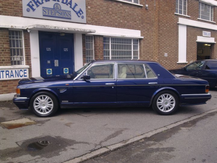 A car parked next to a parking meter - Pistonheads - The image shows a blue limousine parked outside a brick building with white trim around the windows and doors. The building has a sign above the door that reads "Fridel and Stephens." In the background, there is another vehicle, specifically a blue van. The ground appears wet, suggesting it might have rained recently. The car's shiny surface reflects some of the surroundings, enhancing its prominent position in the frame. The overall scene suggests a professional or business environment.