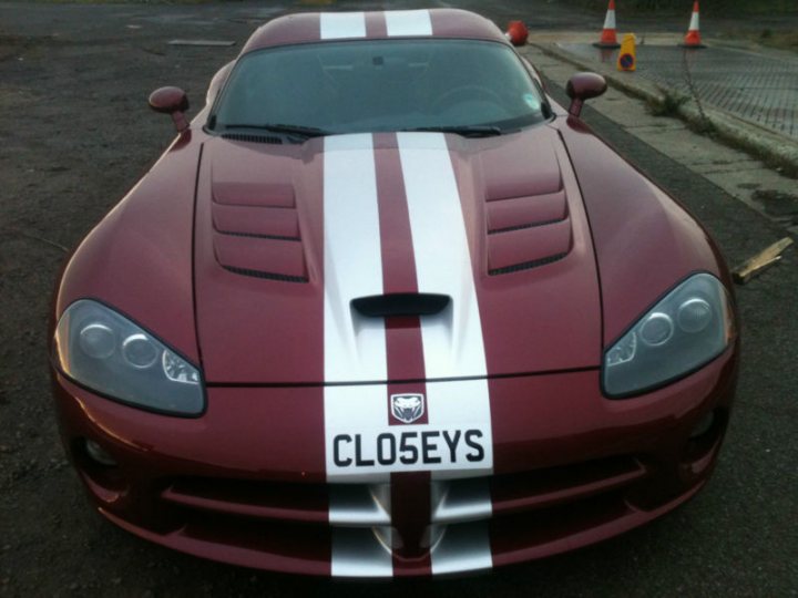 Anyone know this one on ebay?.... - Page 1 - Vipers - PistonHeads - This image features a sleek sports car with a defining red and white stripe running along the center hood. The car is parked on a road that seems to be near a cliff or a dense green area. In the foreground, there is a clear view of the front grille and the word "CLOSEYS" can be seen, perhaps indicating the name of the vehicle or its owner. On the right side of the image, there are orange cones, which might suggest a construction area or the car being parked in a designated space.