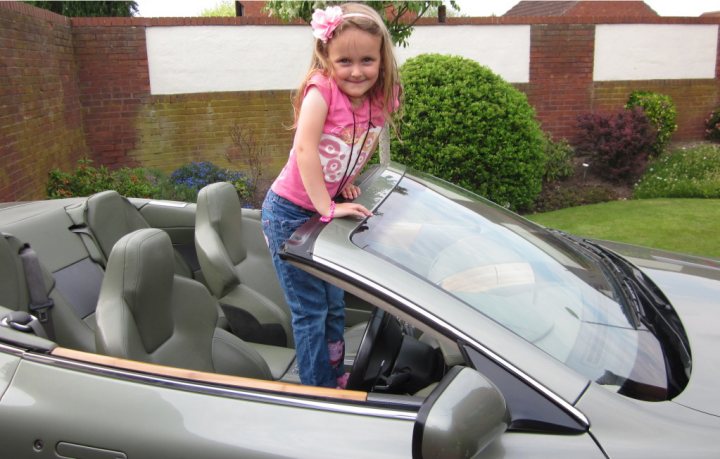 Just had to share this with someone - Page 1 - Aston Martin - PistonHeads - The image depicts a young girl standing in the front seat of a grey convertible car. She is wearing a pink top and jean shorts, with her hand resting on the trunk of the car. The girl appears to be looking towards the camera with a smile. The car is parked in a residential area with a brick fence and various plants visible in the background, suggesting a peaceful suburban setting.