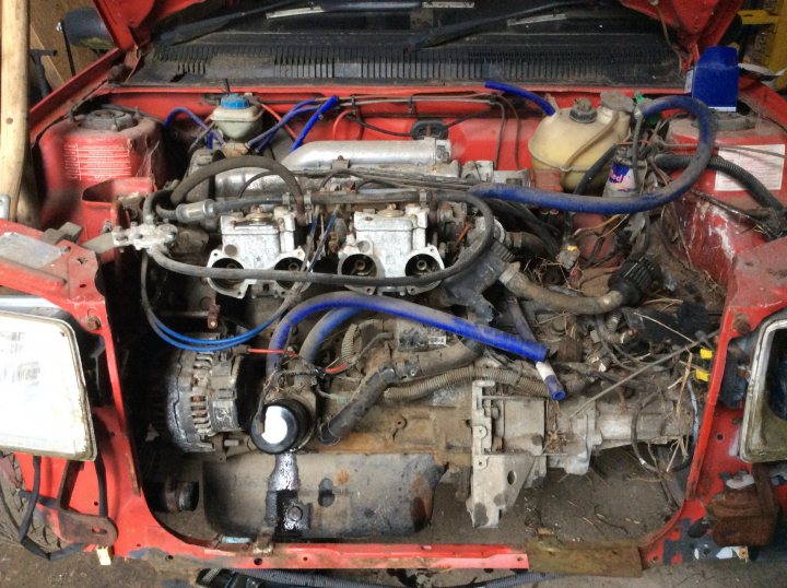 Peugeot 205 gti 1.9 turbo technics ?  - Page 1 - General Gassing - PistonHeads - The image shows a red vehicle that appears to be in the process of being disassembled or repaired. The hood is open, revealing the engine and its components. Various parts are scattered around the vehicle, indicating that work has been done or is ongoing. The setting seems to be an outdoor environment, possibly a garage or a similar workspace. There's no text visible in the image.