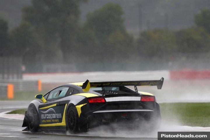 People still looking ? - Page 1 - Gallardo/Huracan - PistonHeads - The image depicts a high-performance sports car, wet from recent rain, taking a curve on a racetrack. The car is predominantly black, adorned with stripes of various colors, including green, white, and yellow. The rear wing of the car is raised, indicating a racing or high-speed mode setting. The track itself is obscured by a light mist, adding to the dynamic, high-speed atmosphere of the scene. The tail-lights of the car are clearly visible, emphasizing its high-performance capabilities. The image captures the car's movement in a way that suggests speed and agility.