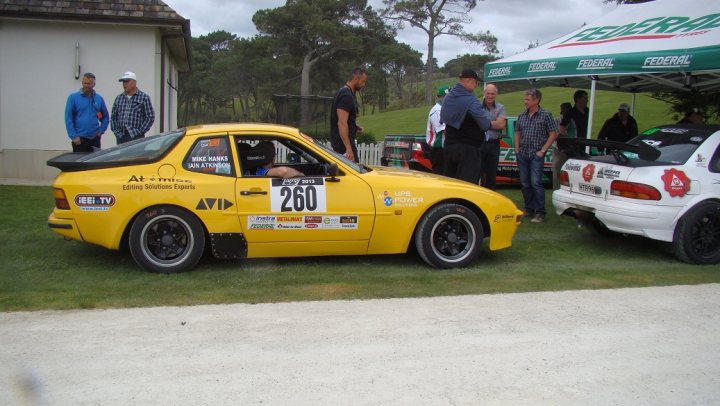 Targa 2013 - Page 1 - New Zealand - PistonHeads - This image captures a lively scene at a classic car show. The cars are arranged in rows, with two black and yellow ones on the left and a yellow one with the number "260" on the right. Several people, some of whom are wearing plaid shirts, are milling about the area, discussing the cars and enjoying the camaraderie. Amidst the grass, a green umbrella pops up, possibly an accessory to one of the cars. The atmosphere is jovial, with a casual and relaxed vibe.