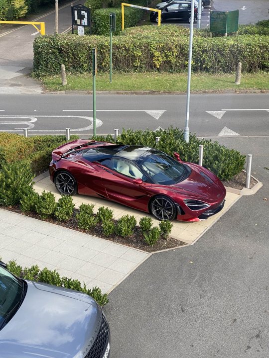 Pick up day for my 720s Volcano Red - Page 1 - McLaren - PistonHeads UK - The image shows a luxurious red sports car parked in a driveway. It's an open-top model with two doors, one for the driver and one for the passenger. The car is positioned in front of a hedge and is adjacent to a sidewalk. There are no visible texts or distinctive markings on the vehicle. In the background, there are other cars parked along the street, indicating that this might be a residential area with parking spaces.