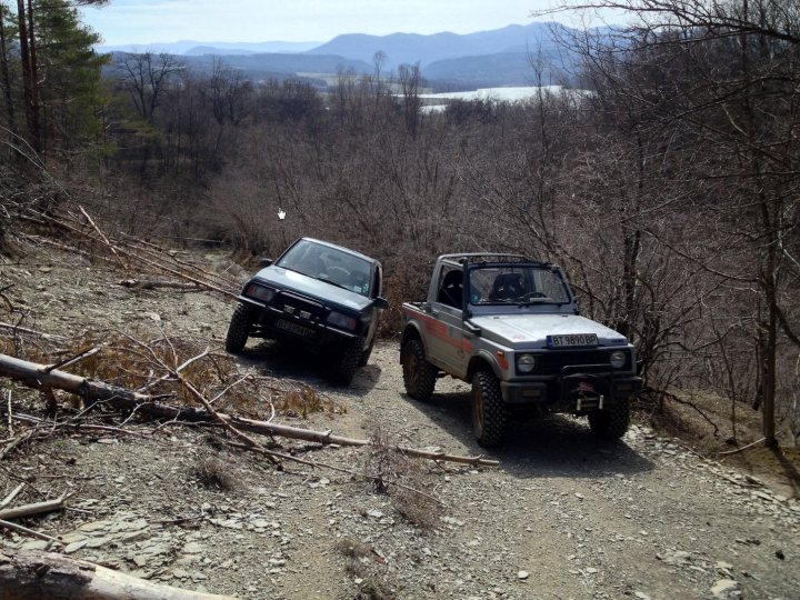 Bit of off-roading, and a bit of camping. - Page 1 - Off Road - PistonHeads - The image depicts a scenic off-road battle between two vehicles on a rugged mountain road. Two four-wheel drive SUVs, one black and the other white, are head-to-head on a dirt trail, their wheels side by side. The terrain is rocky and debris-strewn, suggesting a rugged off-road trail with patches of grass and fallen branches. The backdrop features a blue sky and snow-covered mountains. The parked cars on the side of the road indicate a pause in the race or confrontation between the drivers.