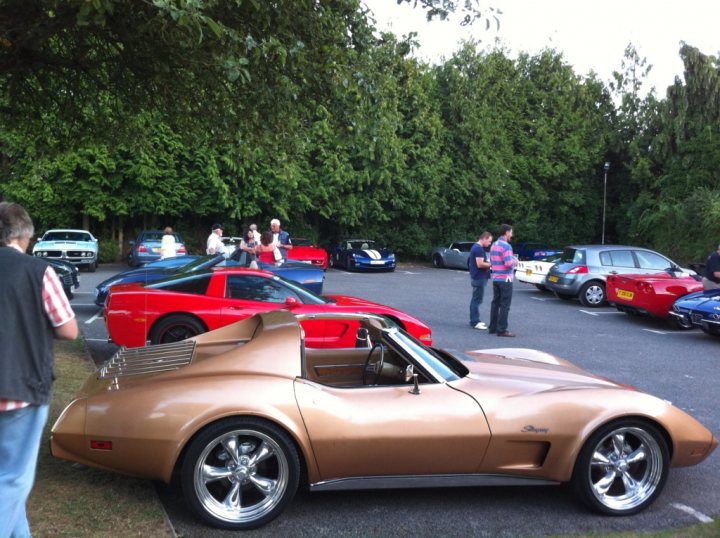 Night July Valley Thames Pistonheads Wednesday - The image captures a gathering of car enthusiasts. Various sports cars, painted in vibrant colors, dominate the scene. They are parked haphazardly around an open area, under the watchful gaze of evergreen trees in the background. Amidst the display of mechanical prowess, a group of people, visibly engrossed in conversation, adds a lively human element to the scene. Their casual attire contrasts with the glossy finishes of the cars, hinting at the relaxed and friendly atmosphere of the event.