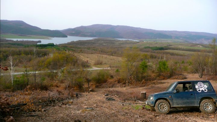 Couple of pics from today... - Page 1 - Off Road - PistonHeads - The image captures a serene outdoor setting featuring a blue truck surrounded by a dense forest. The truck, parked in the middle of the woods, has a skull decal on the back window. Its windows appear to be slightly rolled down. Behind the truck, rolling hills and a river running through them create a picturesque landscape. In the background, scattered buildings can be seen, suggesting that this scene is near a populated area. The atmosphere is tranquil and secluded, evoking a sense of solitude and adventure.