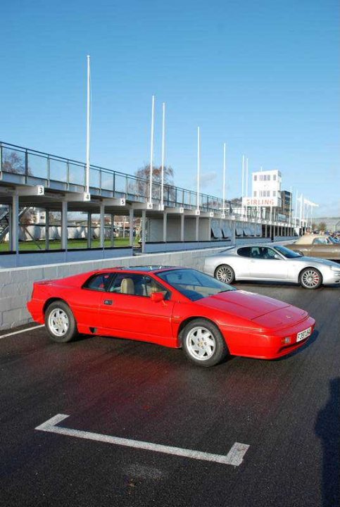 Breakfast Goodwood Pistonheads December - The image shows a red sports car parked in a parking lot. The car is positioned at an angle to a white line marking a parking space. In the background, another red car is visible, parked further back. Beyond the parking lot, there are white pillars and structures that suggest a public or commercial setting, possibly with a sports or racing theme, given the presence of poles and what appears to be a racetrack in the distance. The sky is clear, indicating fair weather. The cars itself possess a sleek, luxurious appearance, which, together with the speed track in the background, suggests a high-performance context.