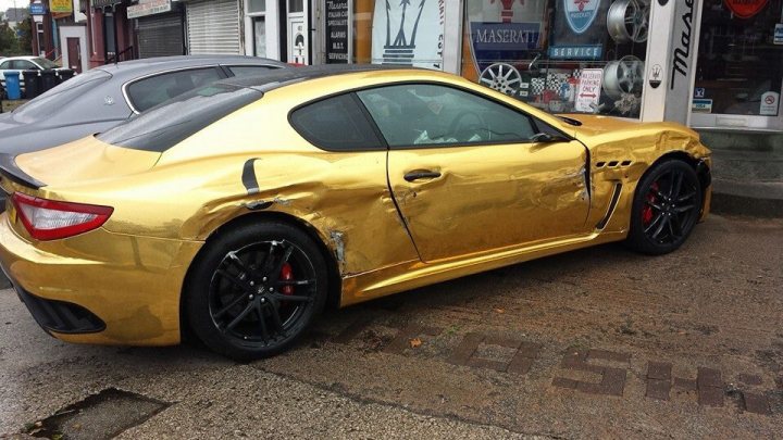 Badly modified cars thread Mk2 - Page 35 - General Gassing - PistonHeads - The image shows a golden sedan parked on a street. The car has a prominent dent and appears to be damaged. It sits next to a building where various signs and a tire rack are visible. There are other cars parked in the vicinity, but this gold car is the main focus of the image due to its shiny color and the noticeable damage. The street scene suggests an urban setting with businesses nearby.