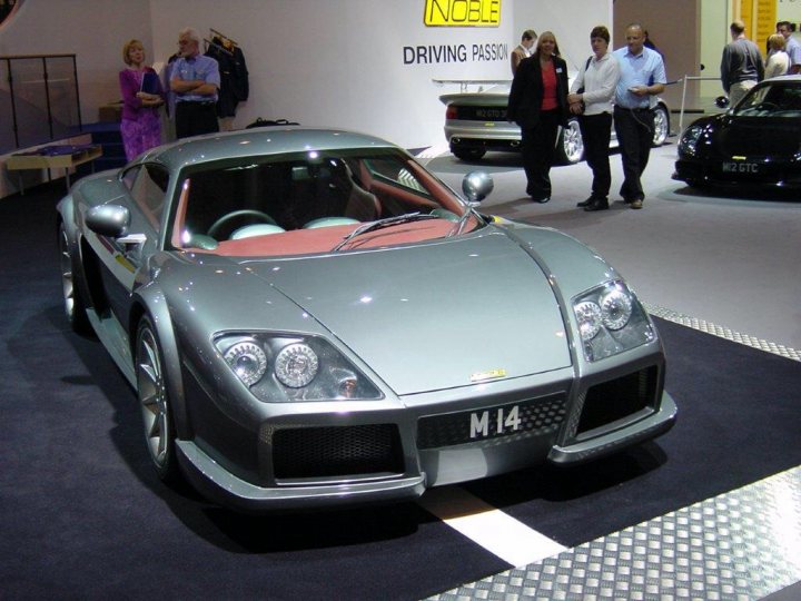 A car that is parked in a parking lot - Pistonheads - The image captures a scene at a car show where a sleek gray sports car is prominently displayed on a gray carpet. The car is positioned inside a showroom, and behind it, multiple onlookers are standing, observing it with interest. The showroom itself appears spacious, allowing ample room for attendees to navigate. A notable detail is the word "NOBLE" prominently displayed in the background, possibly indicating the name of the showroom or the event.