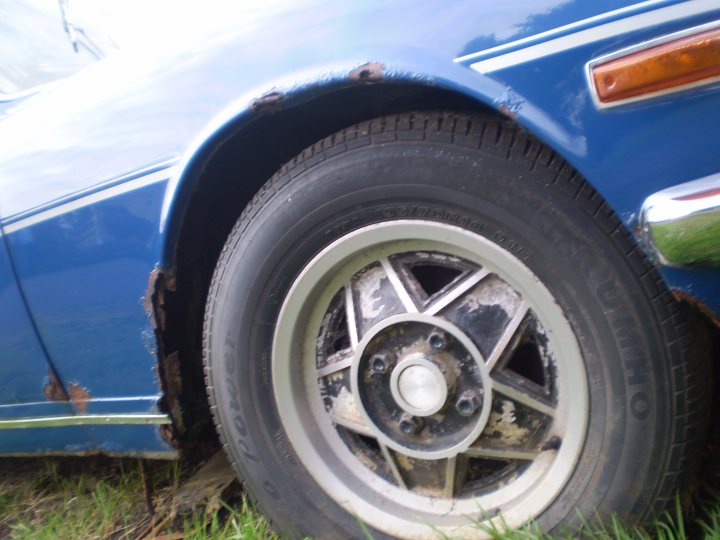 Scrap the Stagg, or give it a new home? - Page 1 - Triumph - PistonHeads - The image shows a close-up of a car's front right tire. The tire has visible signs of wear and damage. The hubcap appears to be silver with a five-pointed star design. The spokes of the wheel are visible, and the centerpiece is round with bolts. The vehicle seems to be parked on a grassy area. The overall condition of the tire suggests it could have been used or neglected for a considerable period.