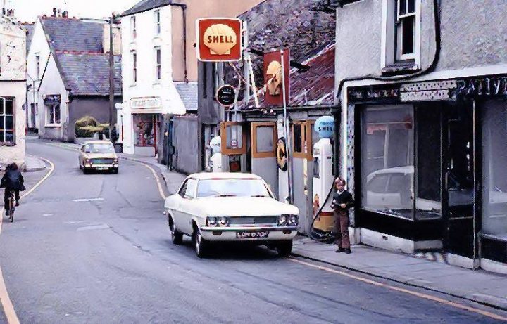 The Humer Unbeam Interesting Filling Stations Thread - Page 46 - General Gassing - PistonHeads - The image presents a scene from the past, featuring a small town street. Cars are parked on both sides of the road, with a person standing near one. A red sign is visible in the background, adding to the urban setting. Buildings line the street, providing a backdrop for this snapshot of everyday life. The overall atmosphere suggests a peaceful day in this quiet town.