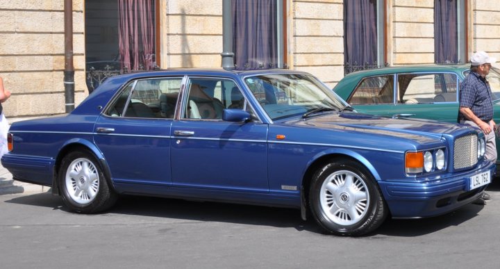 Buy a Bentley for 8K and people think you're a millionaire? - Page 1 - Rolls Royce & Bentley - PistonHeads - The image showcases a classic Bentley car, parked in front of traditional townships. The car is vibrant and well-maintained, featuring a classic design with a distinctive grille and large headlights. There are two people standing on the sidewalk, appearing to observe the car or its surroundings. The building behind them has a European architectural style, with multiple windows and what appears to be a balcony.